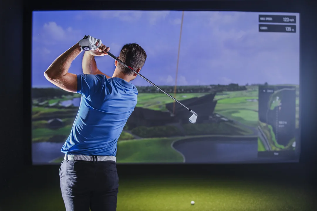 Golfer mid-swing in a Trackman simulator bay with the course on screen
